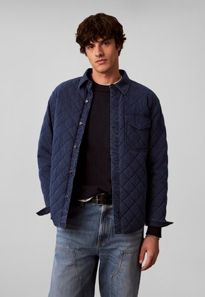 Junger Mann mit kurzen lockigen Haaren, der eine marineblaue gesteppte Jacke über einem schwarzen Pullover und hellblauen Jeans vor einem schlichten grauen Hintergrund trägt.
