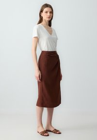 Top bianco a maniche corte con scollatura a V abbinato a una gonna drappeggiata marrone, lunga fino al ginocchio. Il modello indossa sandali marroni con cinturini. Tessuto liscio e casual.
