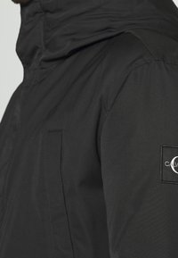 Czarna pikowana kurtka z wysokim kołnierzem, zamkiem z przodu i charakterystyczną naszywką z logo na lewym ramieniu. Gładka, trwała textura materiału.