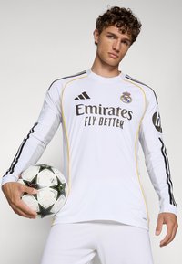 Maglia sportiva a maniche lunghe bianca con dettagli neri e gialli, caratterizzata dal logo "Emirates" e dal testo "Fly Better". Modello che tiene un pallone da calcio.