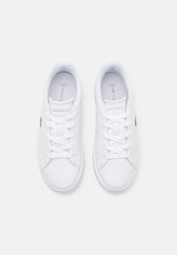 Hvite sneakers laget av glatt syntetisk materiale med flat snøring, og med en liten grønn Lacoste-logo på siden.
