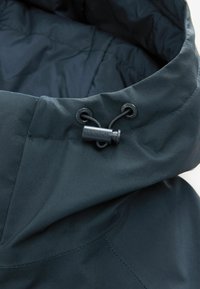 Gulerul jachetei de culoare teal închis este realizat dintr-un material neted și umplut; dispune de șnururi ajustabile și un accent de metal negru inscripționat cu "SIGNATURE".