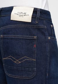 Πίσω τσέπη σκούρου μπλε τζιν με πορτοκαλί ραφές, μικρή κόκκινη λεπτομέρεια "R" και λευκή δερμάτινη ετικέτα με το όνομα «REPLAY DENIM GOODS MFG.CO».