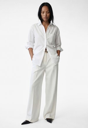 Kvinde iført løs hvid skjorte med knapper og op-rullede ærmer, vidde hvide bukser, sorte spidse sko, stående med hænderne i lommerne.