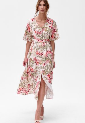 Frau, die in einem weißen Midikleid mit pinken und grünen Blumenmuster geht. Das Kleid hat schwingende Ärmel, einen V-Ausschnitt, einen Taillengürtel und einen Schlitz vorne, dazu trägt sie weiße High Heels.