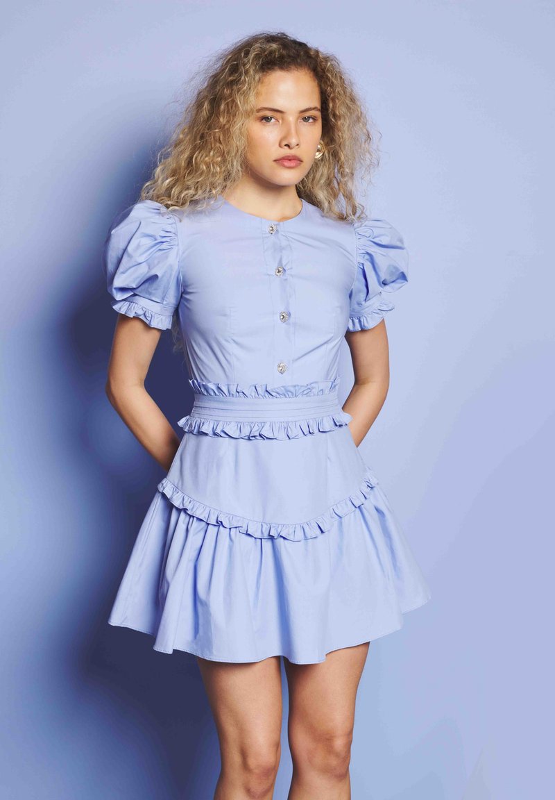 Jauna moteris stovi prieš minkštą mėlyną fono užsklandą, vilkėdama šviesiai mėlyną suknelę su pūkuotomis rankovėmis, mygtukų užsegimu priekyje ir banguotais apvadais.