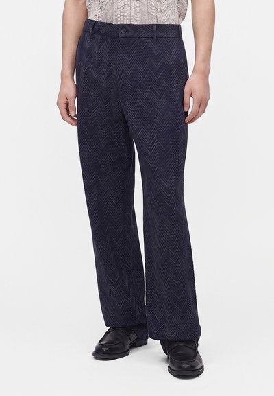 Personne portant un pantalon bleu marine à motif chevron et des chaussures habillées noires, debout contre un fond clair uni.