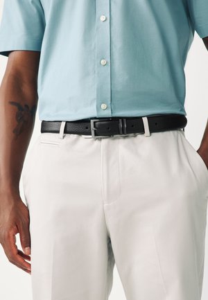 Uomo che indossa una camicia azzurro chiaro a maniche corte infilata in pantaloni bianchi con cintura in pelle nera testurizzata e tatuaggio di un angelo sul braccio sinistro.