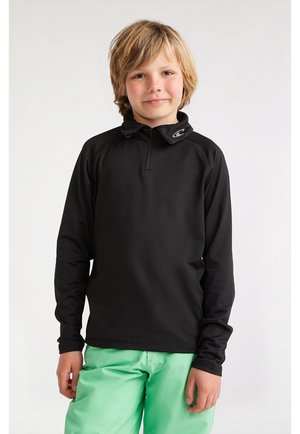 Une chemise noire à manches longues avec une fermeture éclair quart et un col montant, arborant un petit logo sur le col, portée avec un pantalon vert clair.