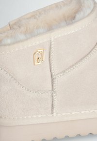 Stivale in camoscio beige con fodera morbida, cuciture decorative e accento del logo dorato. Texture morbida e design arrotondato con suola piatta.
