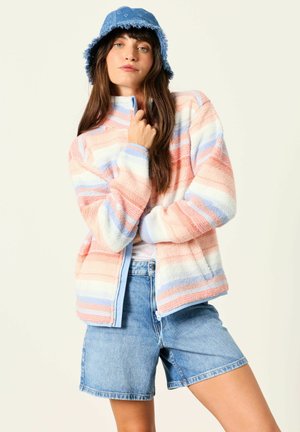 Kvinde iført pastelstribet fleecejakke, denim bucket hat og denimshorts, poserer med hånd, der holder jakke-kraven.