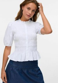 Blouse blanche à manches bouffantes avec smocks texturés, ourlet volanté et fermeture à boutons sur le devant, associée à une jupe en denim foncé.