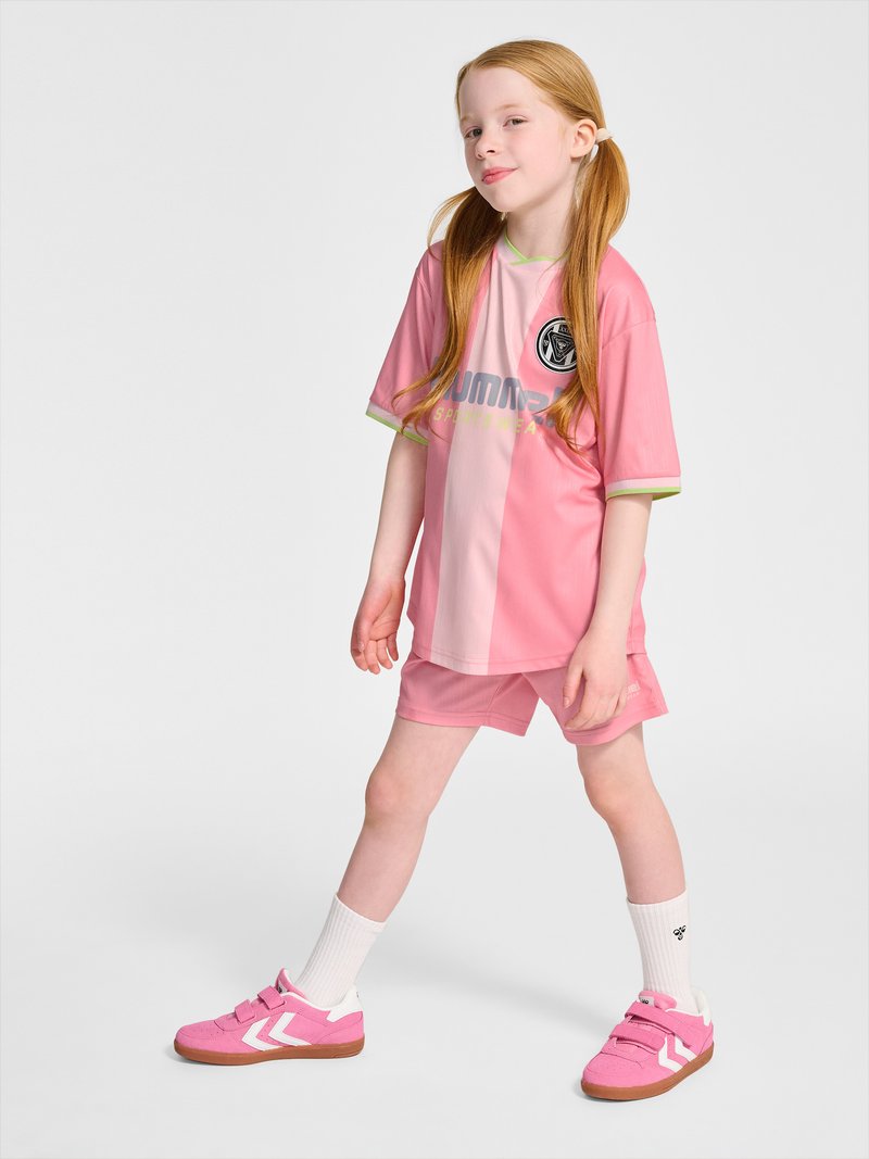 Jong meisje met rood haar in roze sportkleding staat met gespreide benen op een effen witte achtergrond.