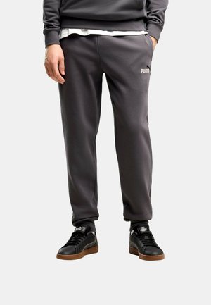 Homme portant un pantalon de jogging Puma gris foncé et des baskets Puma noires avec des semelles marron, debout, une main dans la poche, sur un fond clair.
