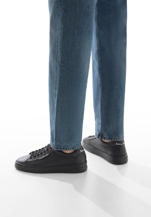 Schwarze Leder-Sneaker mit strukturierten Akzenten, runder Zehenpartie und flacher Gummisohle, getragen mit hellblauen geraden Jeans.