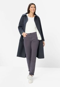 Lang mørkegrå uldfrakke med hvid foring, båret over en hvid top og mørkelilla bukser. Fremhævet med hvide sneakers.