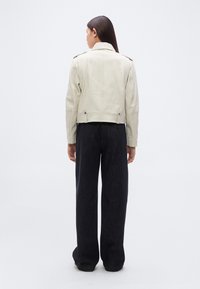 Μπεζ δερμάτινο μπουφάν με κοντό μήκος, διακοσμημένο με επιταχυντές στους ώμους και εφαρμοστή μέση, συνδυασμένο με φαρδιά μαύρη παντελόνα.