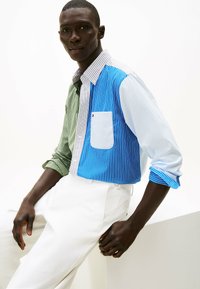 Koszula w paski w wielu kolorach, z zielonymi, niebieskimi i białymi sekcjami oraz kieszenią na piersi, zestawiona z białymi spodniami. Gładka tkanina, dopasowany krój.