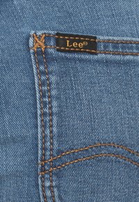 Jeans de denim azul con costuras naranjas contrastantes, un bolsillo con una etiqueta negra que dice "Lee" y una textura suave.