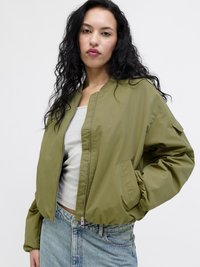 Mujer con cabello largo y rizado, lleva una chaqueta bomber verde oliva sobre una camiseta blanca y vaqueros azul claro, de pie frente a un fondo liso.