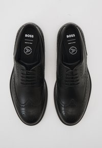 Juoda odinė klasikinė brogué stiliaus batų pora su raišteliais, viduje označiu „BOSS“ ir „Pagaminta Italijoje“, rodoma iš viršaus ant balto fono.