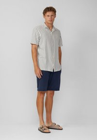 Korte mouwen gestreept shirt in crème en blauw, gecombineerd met marineblauwe shorts en bruine sandaal met twee banden. Casual zomeroutfit.