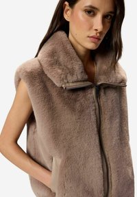 Telovnik s kratkimi rokavi iz mehke, svetlo rjave umetne kože z visokim, mehkim ovratnikom. Ima sprednjo zadrgo in stranske žepe z usnjenim robom.