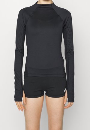 Osoba nosící černé přiléhavé sportovní tričko s dlouhým rukávem a otvory na palec a černé šortky Adidas na jednobarevném pozadí.