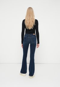 Jeans acampanados de mezclilla azul oscuro con una altura de cintura media, que cuentan con dos bolsillos traseros, costuras sutiles y una etiqueta de cuero en la cinturilla.