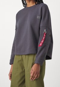 Šedá mikina s kulatým výstřihem a žebrovanými manžetami, zdobená kapsou na zip s červenou štítkem, nošená přes olivově zelené kalhoty.