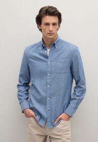 Camicia azzurro chiaro con bottoni frontali, maniche lunghe, taschino sul petto e colletto, realizzata in tessuto morbido. Indossata con pantaloni beige.