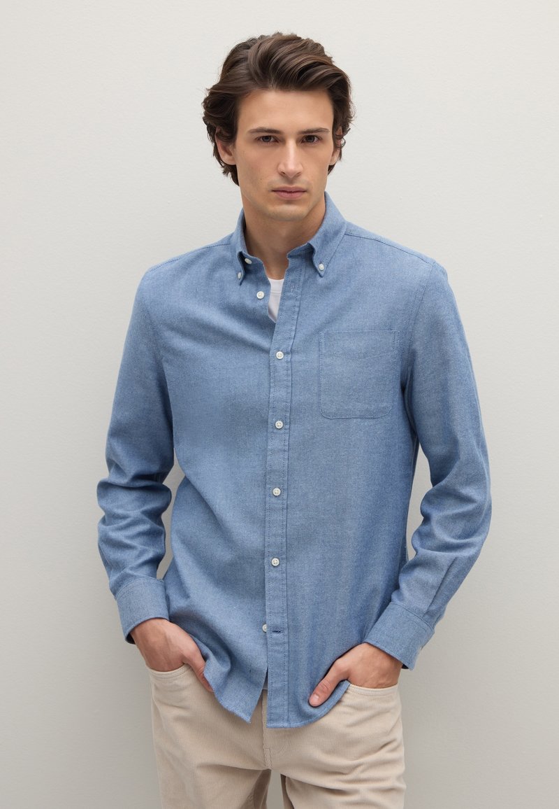 Camicia azzurro chiaro con bottoni frontali, maniche lunghe, taschino sul petto e colletto, realizzata in tessuto morbido. Indossata con pantaloni beige.