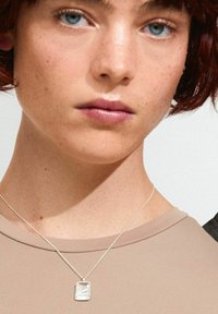 Collar de plata con un colgante en forma rectangular irregular, llevado sobre una camisa marrón claro, con una cadena delicada.