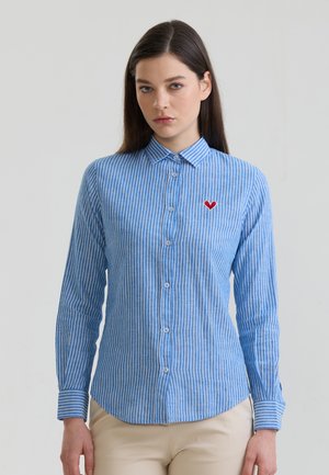 Kobieta o długich ciemnych włosach, ubrana w niebiesko-białą paskowaną koszulę na guziki z małym czerwonym sercem na lewej piersi.