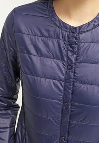Navy puffer jacket featuring horizontal quilted sections, round neckline, and button closures, made from shiny, lightweight material.