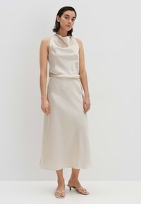 Robe satin beige avec un décolleté drapé, sans manches, et une jupe mi-longue avec une fente sur le côté. Comprend des sandales plates minimalistes.