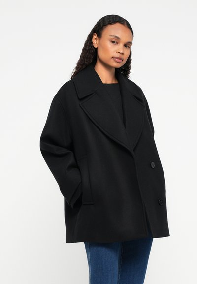 Mulher com cabelo longo e encaracolado, vestindo um casaco preto de duplo peito largo, top preto e jeans azuis, de pé com as mãos nos bolsos do casaco.