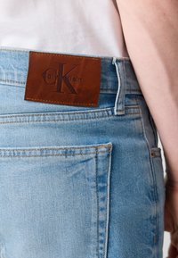 Svijetloplave traperice s smeškim kožnim Calvin Klein pečatom na struku, sa detaljima šivanja i standardnim dizajnom s pet džepova.