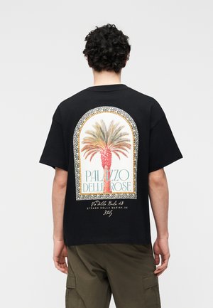 Homme portant un t-shirt noir avec un palmier coloré et le texte "Palazzo Delle Rose" encadré par un motif en mosaïque au dos.