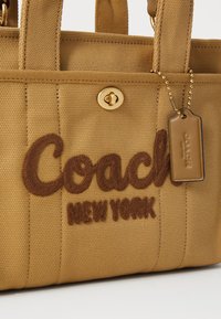 Sac fourre-tout Coach en toile beige avec logo brodé marron, fermeture à loquet doré et étiquette en cuir sur chaîne à billes dorée.