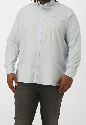 Homme portant une chemise bleu clair boutonnée et un pantalon gris foncé, debout les mains près des hanches sur un fond uni.