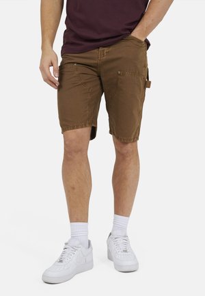 Homme portant un short cargo marron, des chaussettes blanches et des baskets blanches, debout avec une main dans la poche sur un fond blanc.