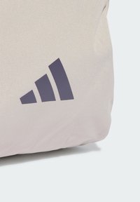 Borsa di tessuto beige chiaro con superficie testurizzata, caratterizzata da un logo Adidas viola scuro con tre strisce diagonali su un lato.