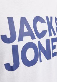 Tricou alb din bumbac, având litere mari albastre, imprimate, care formează cuvântul "JACKS JONES". Materialul are o textură netedă și un design clasic cu guler rotund.