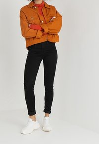 Chaqueta de mezclilla naranja, suéter de cuello alto rojo, jeans ajustados negros y zapatillas blancas. La chaqueta tiene dos bolsillos frontales y puños abotonados.