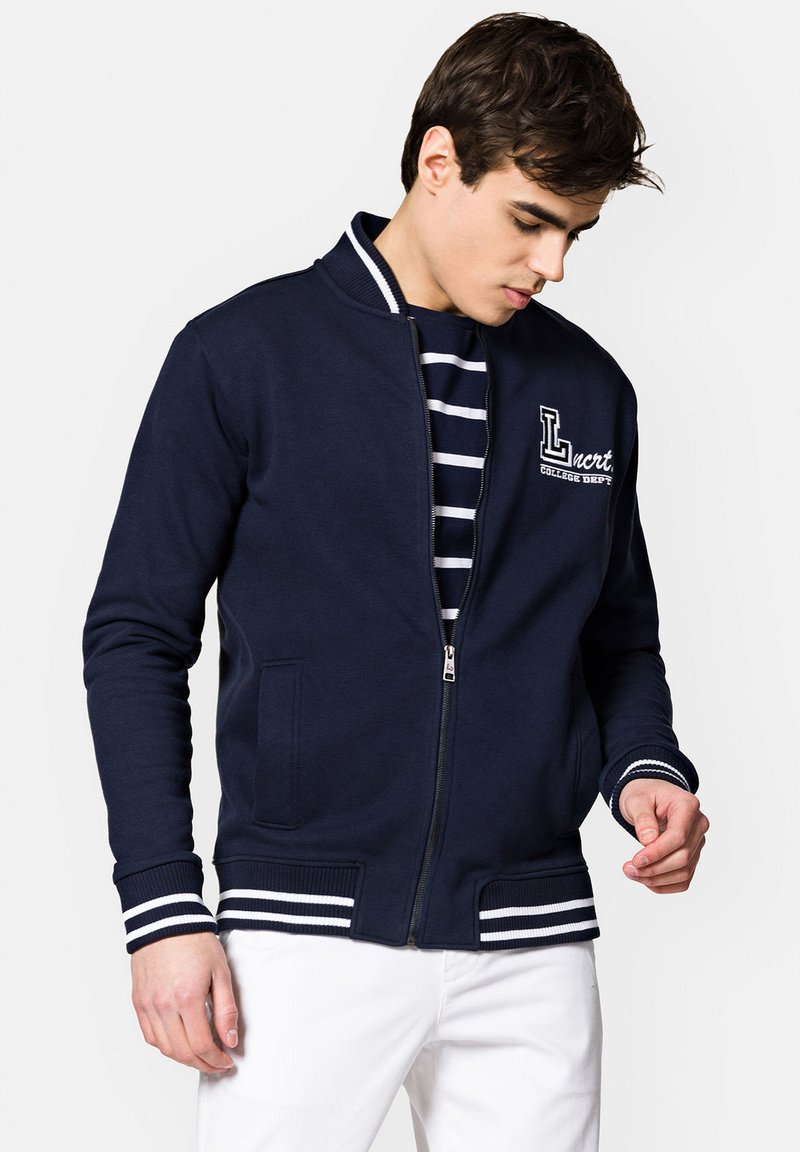 Granatowa bomberka zapinana na zamek z prążkowanymi mankietami i paskiem w talii, z białymi paskami i logo na lewej piersi. Wykonana z miękkiego materiału.