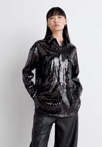 Schwarzes Pailletten-Hemd mit Knopfleiste, Kragen, langen Ärmeln und Fronttaschen. Der Stoff hat eine glänzende und reflektierende Textur, die einen gemusterten Effekt erzeugt.