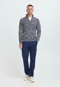 Hombre de pie vestido con un suéter con cremallera gris de estampado, pantalones cargo azul marino y zapatillas blancas contra un fondo blanco simple.