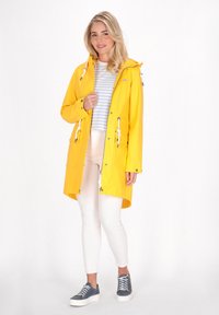 Rumena vodoodporna dežna jakna s kapuco, sprednjim zadrgo, vrvicami in dvema žepoma. Nosijo jo čez svetlo črtasto srajco in bele hlače.
