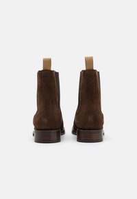Bottines en daim marron avec des panneaux extensibles et des tirants en tan. Elles présentent un talon en bois et une silhouette simple et épurée.